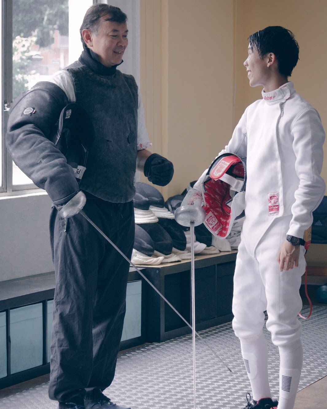 Coach Jeffrey Lopez and Gordon Ho in fencing kit, talking and laughing between bouts
